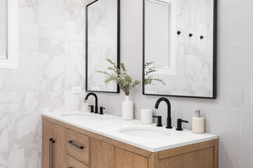 A bathroom with marble and stacked vertical subway tiles, a white oak vanity cabinet, black framed square mirrors and faucets, and marble countertop. - obrazy, fototapety, plakaty