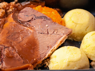 Classic beef dinner meal with slice of meat, mashed potatoes, gravy and turnip in dark plastic container. Close up.