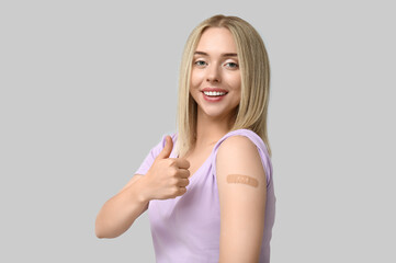 Young woman with medical patch on arm showing thumb-up against grey background. Vaccination concept