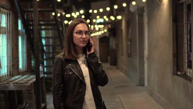 A young woman talks on the phone on a narrow street in the evening. A girl with glasses and a leather jacket on a cozy street.