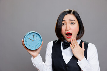 Shocked overslept asian waitress holding alarm clock and looking at camera while covering open mouth. Young anxious woman receptionist showing time while running late portrait