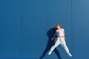 Joyful young European blond girl jumping in a white T-shirt and white sweatpants with pink heart-shaped glasses against a blue wall outside