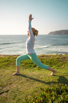 Pensioner woman in warrior yoga pose on coast. Fitness practice of female, who stand barefoot on green grass near sea shore. Side view of mature woman in sportswear doing yoga workout on nature