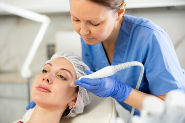 Adult woman cosmetologist performs hardware facial procedure to young woman patient