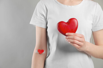 Blood donation concept. Woman with adhesive plaster on arm holding red heart against grey...