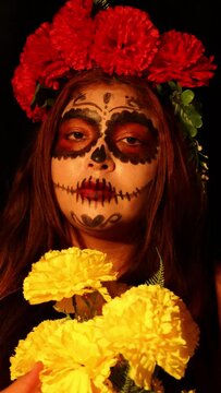 Woman dressed as catrina holding a bouquet of yellow flowers in her hands with day of the dead makeup on a dark background