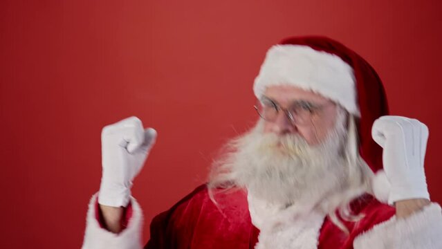 Medium close-up shot of Santa Claus in red suit, pointed hat and glasses, with grey hair, beard and moustache dancing energetically with batman moves on plain red background. Template, copy space