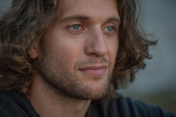 Close-up of young adult handsome man with long curly hair and stubble looks forward outdoor 4K