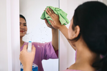 Rear view of pretty Asian woman cleaning mirror with rag and spraying detergent 