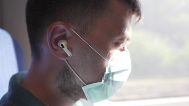 Topic travel by public transport during coronavirus pandemic. A passenger of a train sits by the window and uses white headphones wearing a protective mask against infection covid 19.
