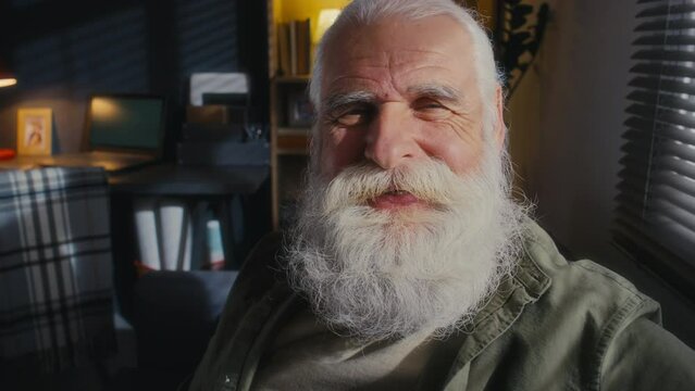 Close-up portrait shot of pensive senior male widower with grey hair, thick beard and moustache sitting by window at home, looking outside, then at camera and smiling