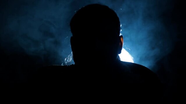 Close Silhouette shot of a female sitting in front of a spotlight wearing a jacket in a smoky dark mystic environment.