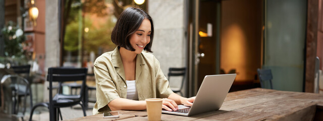 Young asian woman, digital nomad working remotely from a cafe, drinking coffee and using laptop,...