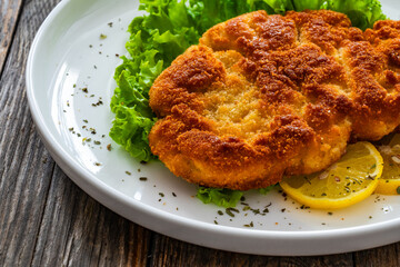 Crispy breaded seared chicken cutlet with lemon slices and fresh vegetables on wooden table
