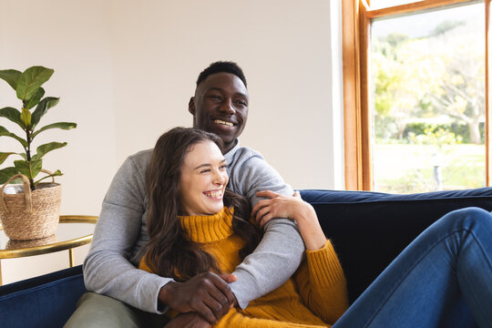 Happy diverse couple sitting on sofa embracing and smiling at home