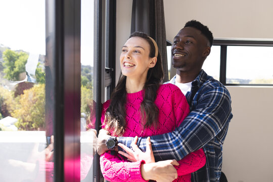 Happy diverse couple embracing and looking through window at home
