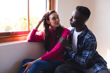 Diverse couple embracing and talking next to window at home