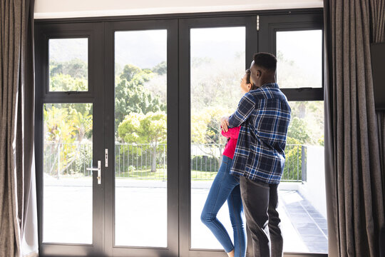 Happy diverse couple embracing and looking through window at home, copy space