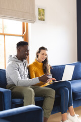 Happy diverse couple sitting on sofa using laptop for online shopping at home