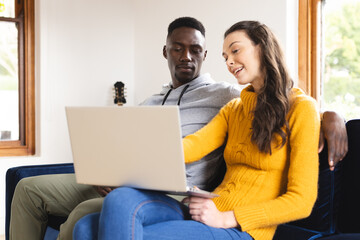 Happy diverse couple sitting on sofa using laptop at home