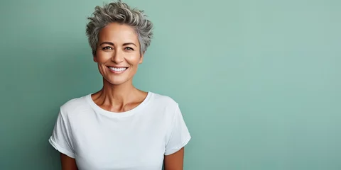 Fotobehang Beautiful mature woman in her fifties with turquoise background, smiling senior lady in a white t-shirt, studio shot with copy space © leftmade