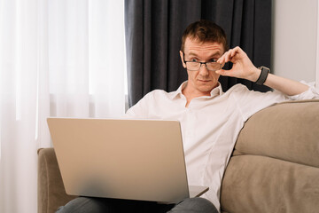 Mature business human watching screen with goggle eyes, adjusting glasses. Man surprised while working on home couch.