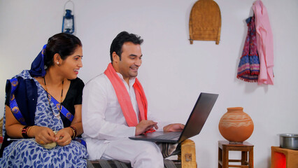 Village scene of a middle-aged couple happily spending time together at home. Handsome Indian villager showing new things to his beautiful wife on a laptop while she is knitting woolen clothes - obrazy, fototapety, plakaty