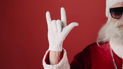 Close-up half portrait shot of hipster Santa Claus with grey beard, in sunglasses, showing horns hand sign, on plain red background. Suitable as template, copy space