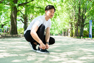 Young Asian sportsman kneeling and tying shoelaces in the park, getting ready to jog outdoors....