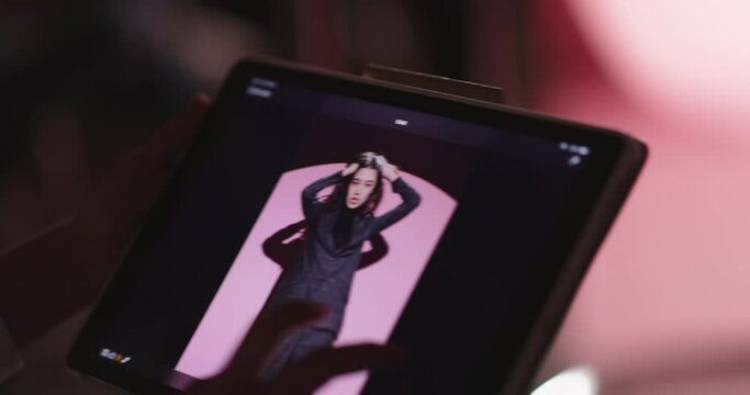 Backstage professional fashion photo shoot. Unknown person uses digital tablet with footage from professional photo shoot. Results checking. Young model in black pantsuit on pink background