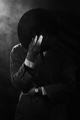 a dark silhouette of a man in a raincoat and a hat in the style of crime noir. Dramatic noir portrait in the style of detectives of the 1950s. There is smoke in the photo - obrazy, fototapety, plakaty