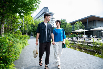 Fototapeta premium A beautiful couple, a man and a woman, are walking happily hand in hand through a green shopping commercial area. They are smiling and chatting. Urban dwellers. Healthy lifestyle.