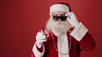 Medium shot of senior man in Santa outfit, hat, with silver chain, beard and moustache drinking whiskey out of tumbler, pulling down sunglasses and winking, on red background. Template, copy space