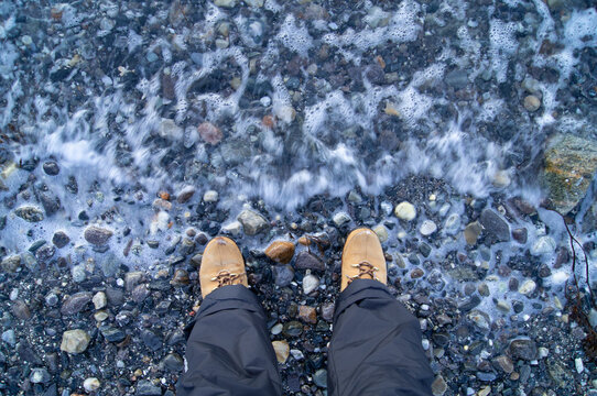Person, human, man, standing at water, in boots. Sea, fjord, lake, winter clothes, boots. Looking, look down, edge, end. Pebels, pair.