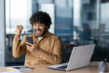 Portrait of hispanic winner at workplace, businessman received online win notification on phone app, man celebrating victory joyfully holding hand up, inside office with working with laptop.