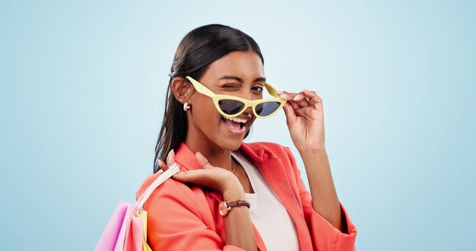 Sale, shopping and portrait of happy woman with bags of clothes, retail product or wink on studio blue background. Fashion, deal and Indian gen z model with sunglasses and offer with store discount