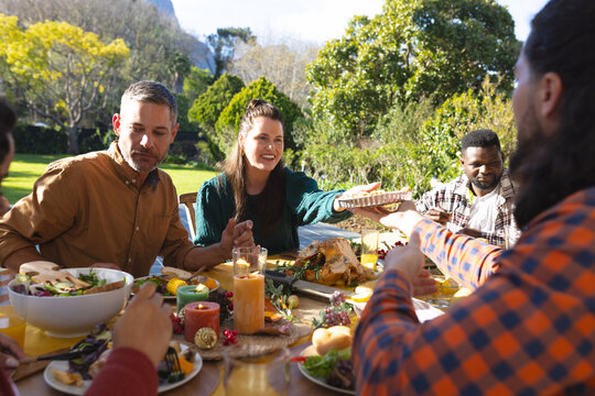 Happy diverse male and female friends eating thanksgiving celebration meal in sunny garden