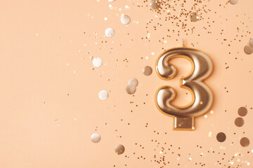 Gold candle in the form of number three on peach background with confetti.