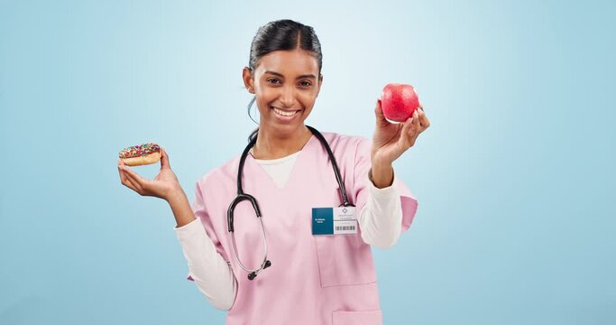 Woman nurse with apple and donut for choice with nutrition, diet or balance in studio. Options, medical and Indian female doctor nutritionist with sweet dessert and fruit isolated by blue background.