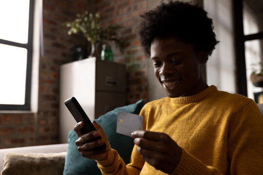 Happy African American Man At Home Holding Credit Card And Using Smartphone To Make Online Payment