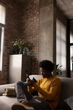 Happy african american man sitting on couch at home using smartphone, copy space