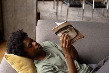 Happy african american man lying on couch reading book wearing under eye masks