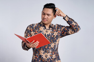 Unhappy young Asian man wearing batik shirt reading study book and scratching his head with...