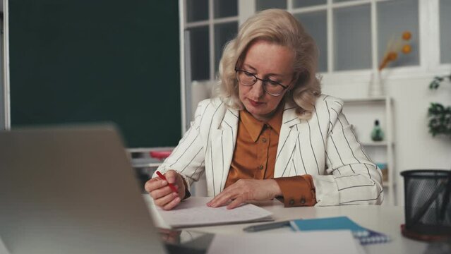 Middle aged teacher grading students papers in classroom, tutor occupation