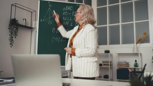 Woman math teacher writing geometry formula on blackboard during online lesson