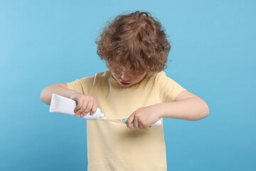 Cute little boy squeezing toothpaste from tube onto electric toothbrush on light blue background