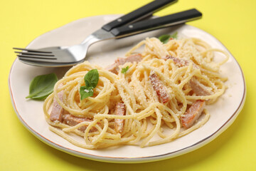 Plate of tasty pasta Carbonara with basil leaves, fork and spoon on yellow background, closeup