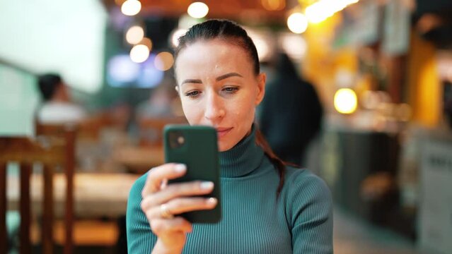 Portrait of beautiful young woman who uses mobile phone and looks in screen, reading text or texting and communicating on social networks. Girl sitting in public place, in shopping mall or restaurant.
