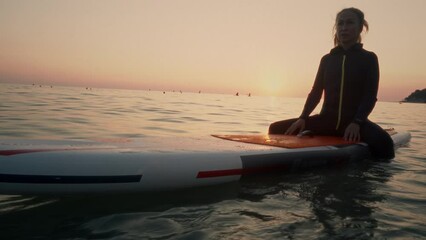 An elderly woman rides paddleboarding in the ocean. Senior female is waiting for a wave on a surfboard. Active old age. Old girl is sitting on a board in the sea at sunset, dressed in a swimming suit