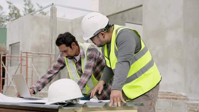 Asian male engineer and construction foreman Wear reflective clothing Wear a safety hat Standing and looking at house from the blueprint on the table. Discuss the progress of the house construction.